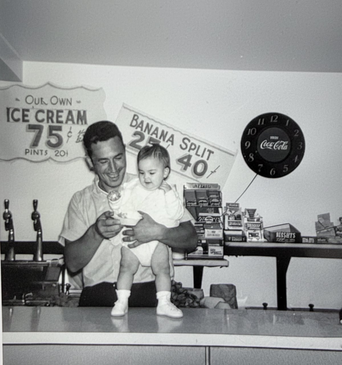 A man holds a baby behind an old-fashioned ice cream counter. Signs advertise ice cream and banana splits. Candy bars and a Coca-Cola clock are on the wall. The scene appears to be from the mid-20th century.