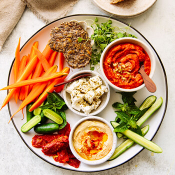 A platter with carrot sticks, cucumber slices, crackers, herbs, sun-dried tomatoes, feta, hummus, and red pepper dip in bowls, with a plate of rustic bread in the background.