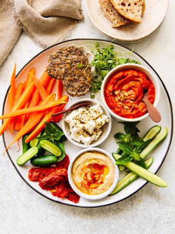 A platter with carrot sticks, cucumber slices, crackers, herbs, sun-dried tomatoes, feta, hummus, and red pepper dip in bowls, with a plate of rustic bread in the background.