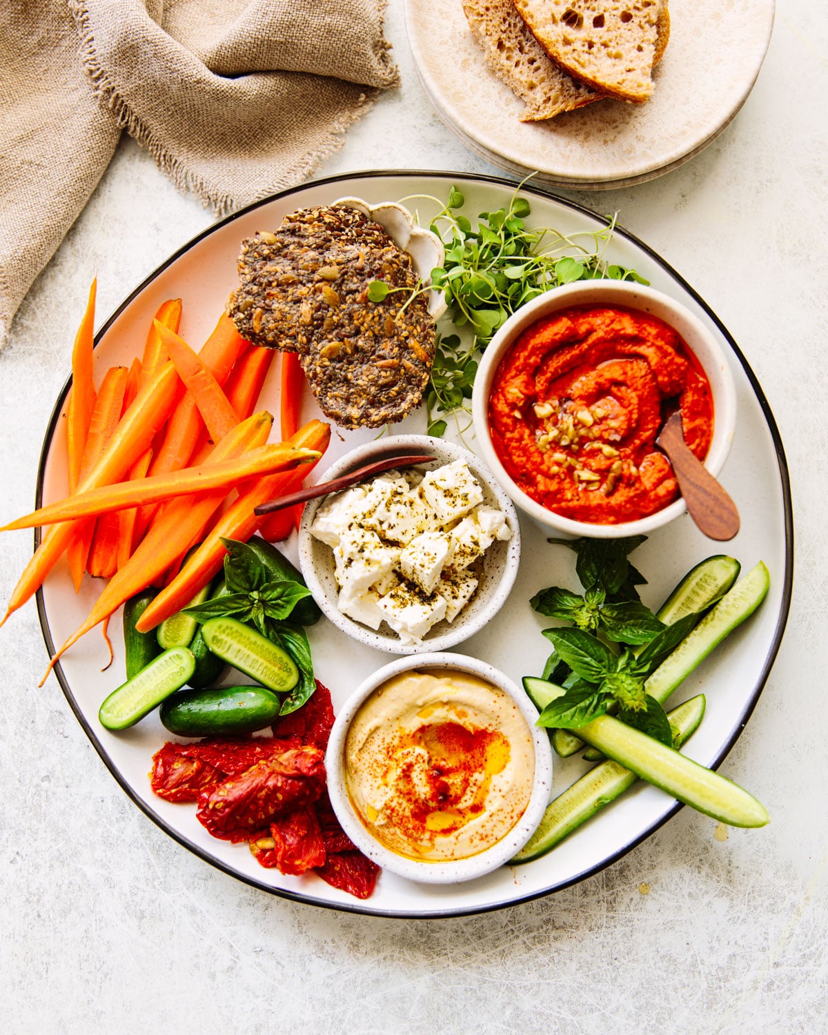 A round platter with three dips — herby feta, whipped hummus, and muhammara — surrounded by carrot and cucumber sticks, sun-dried tomatoes, seeded crackers, and fresh basil and microgreens.
