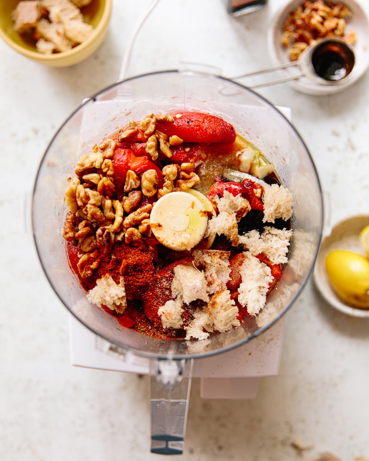 A food processor bowl filled with tomatoes, walnuts, bread pieces, spices, and seasonings sits on a white counter, surrounded by a lemon, a small bowl of walnuts, and a tablespoon of pomegranate molasses.