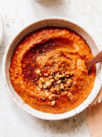 A bowl of red pepper dip garnished with chopped walnuts and spices, with a wooden spoon resting inside. The bowl is on a light textured surface, next to a beige cloth.