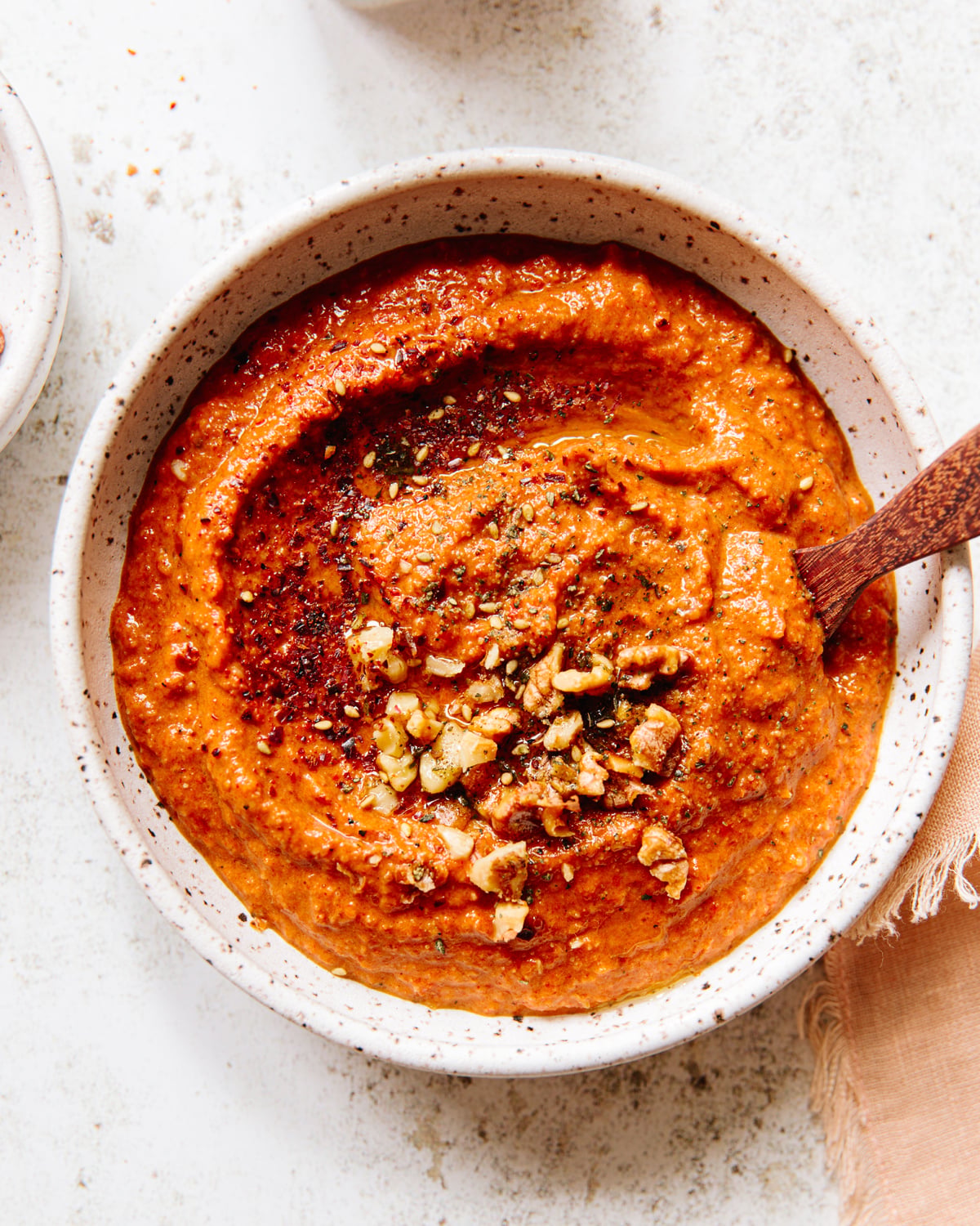 A bowl of red pepper dip garnished with chopped walnuts and spices, with a wooden spoon resting inside. The bowl is on a light textured surface, next to a beige cloth.