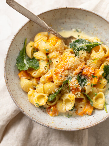 A bowl of creamy pasta shells with kale, roasted chunks of pumpkin or butternut squash, and grated Parmesan cheese, served with a fork on a light-colored plate atop a neutral cloth.