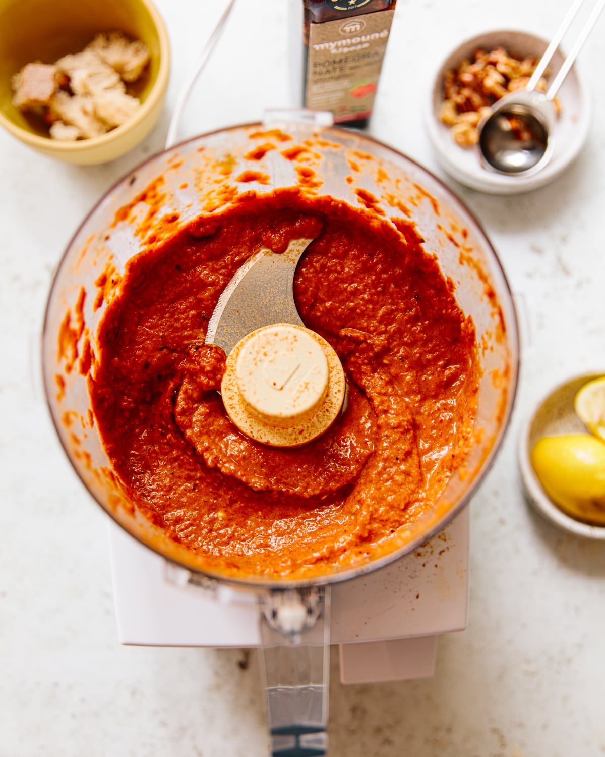 A food processor filled with blended muhammara sauce sits on a counter, surrounded by a yellow bowl, a lemon half, measuring spoons with spices, and a jar in the background.