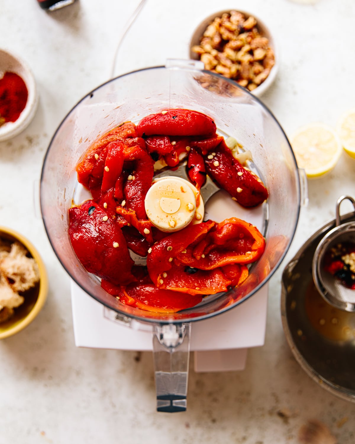 Food processor bowl filled with roasted red peppers and seasonings, surrounded by small bowls of walnuts, lemon halves, brown sugar, and spices on a white countertop.