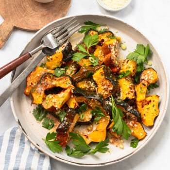 Plate of roasted squash slices garnished with fresh parsley, chopped green onions, and grated cheese, with a fork and knife on the side. Small bowls of crushed red pepper and grated cheese are nearby.