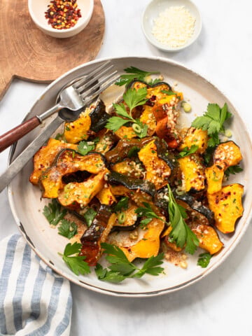 Plate of roasted squash slices garnished with fresh parsley, chopped green onions, and grated cheese, with a fork and knife on the side. Small bowls of crushed red pepper and grated cheese are nearby.