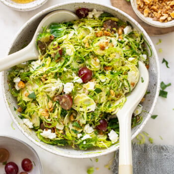 A serving bowl of shaved Brussels sprouts salad with grapes, walnuts, and crumbled goat cheese, plated with serving spoons.