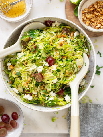 A serving bowl of shaved Brussels sprouts salad with grapes, walnuts, and crumbled goat cheese, plated with serving spoons.