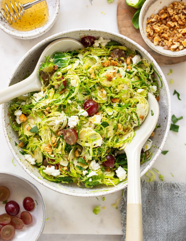 A serving bowl of shaved Brussels sprouts salad with grapes, walnuts, and crumbled goat cheese, plated with serving spoons.